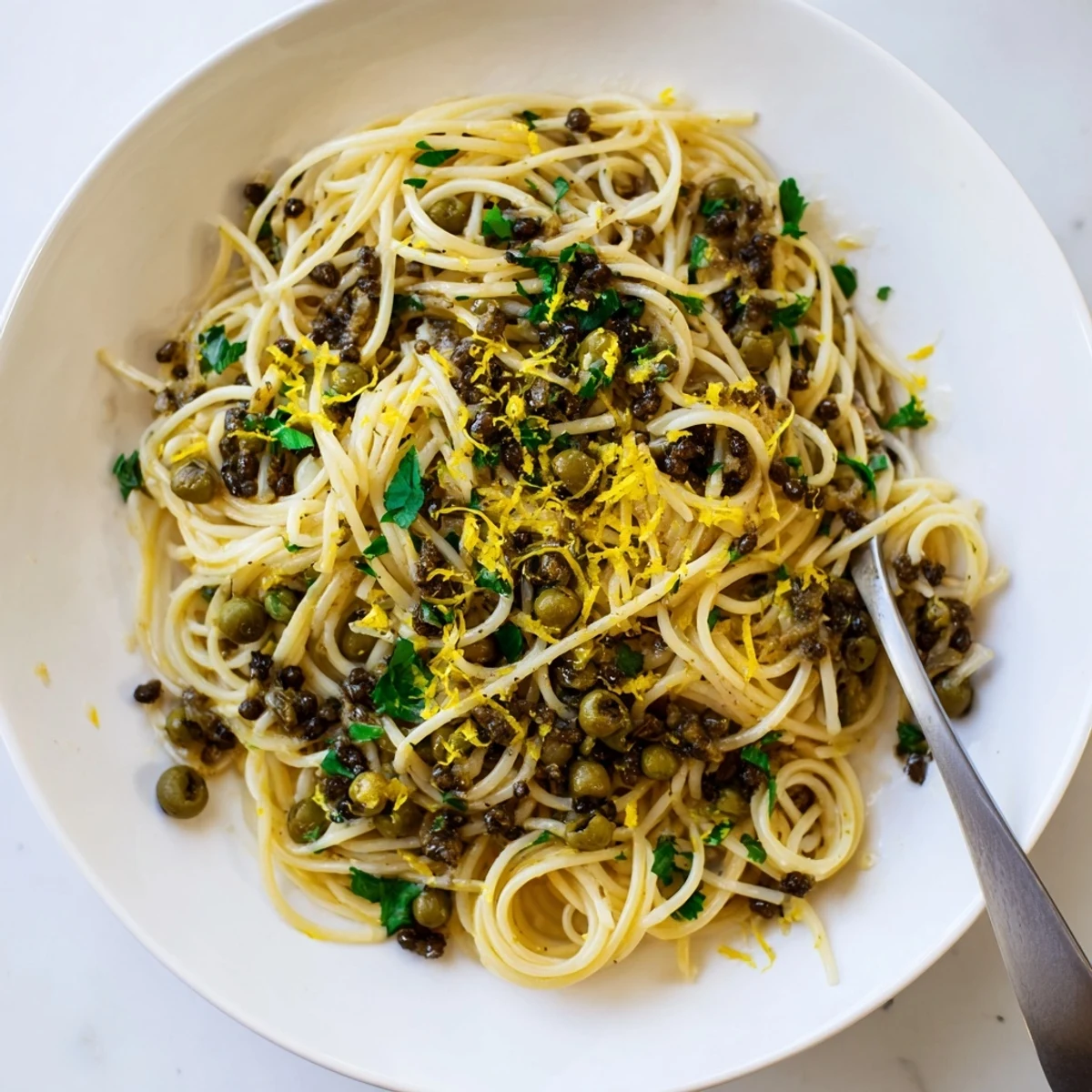 A close-up of Olive Tapenade Pasta twirled on a fork, with chopped parsley and lemon zest garnish.